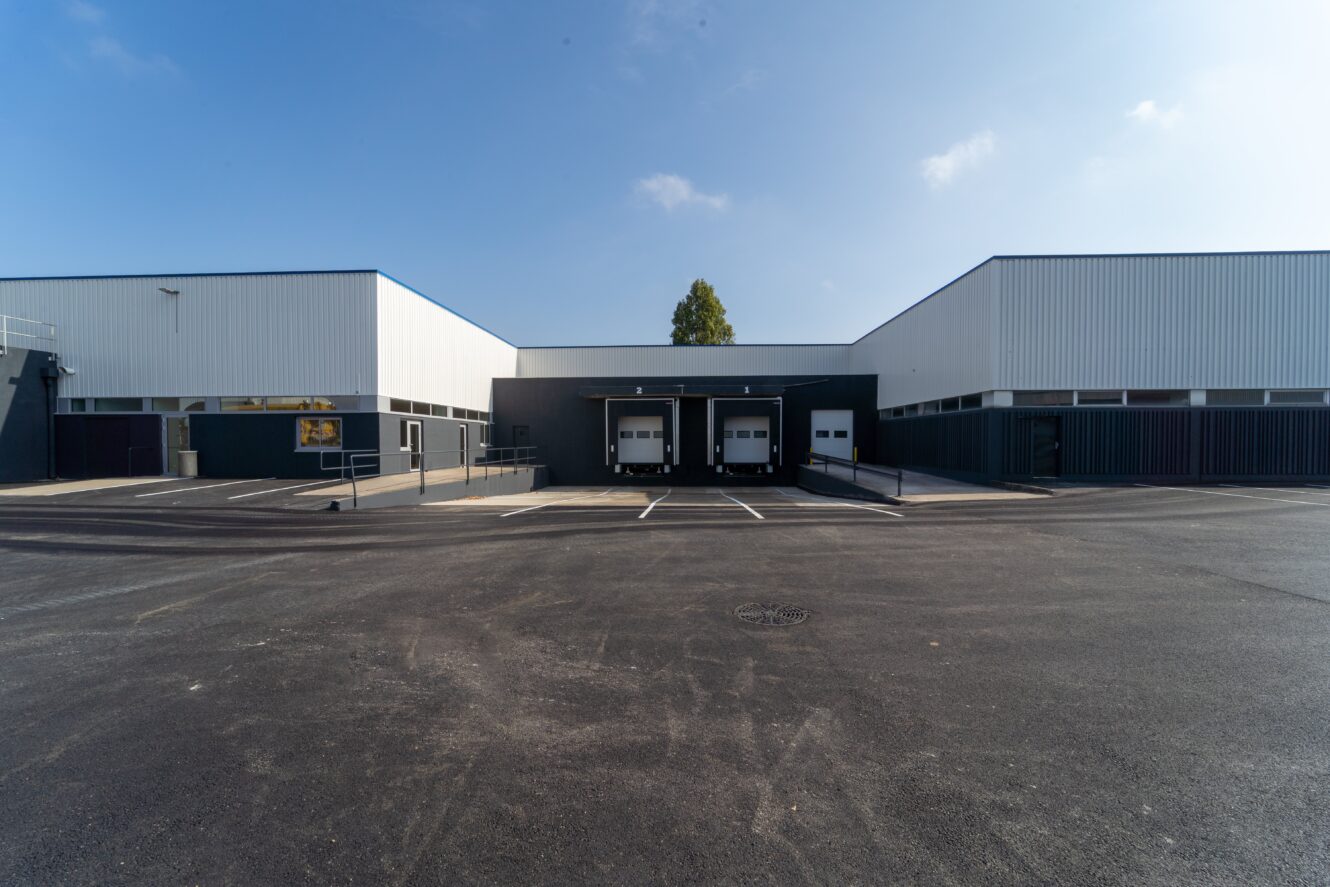 Un grand bâtiment industriel avec trois quais de chargement, des places de parking vides et un ciel bleu clair au-dessus de la tête.