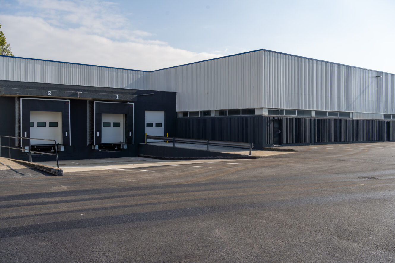Un entrepôt industriel moderne avec deux quais de chargement, un bardage métallique et un terrain bitumé vide à l'avant.