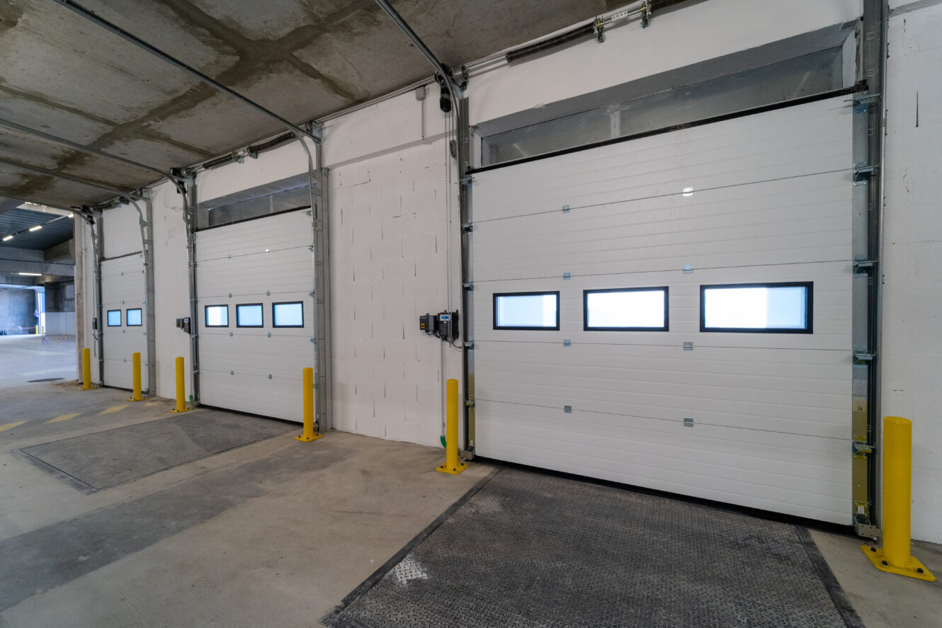 Trois portes de garage industrielles fermées avec de petites fenêtres rectangulaires, des bornes de sécurité jaunes et un sol en béton à l'intérieur d'un entrepôt ou d'un quai de chargement.