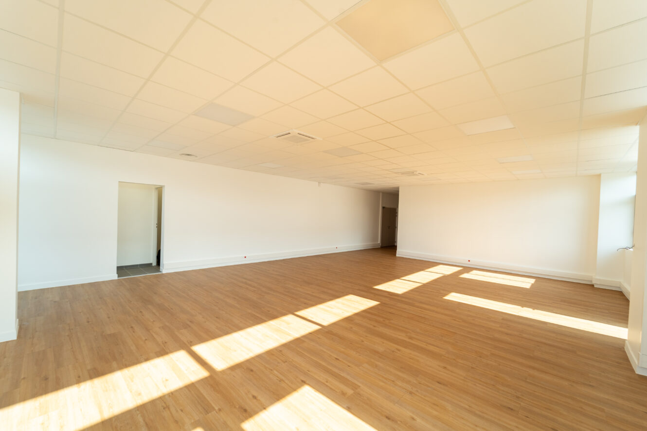 Une pièce vide aux murs blancs, au sol en bois clair et au plafond quadrillé. La lumière du soleil traverse de grandes fenêtres, créant des taches lumineuses sur le sol.