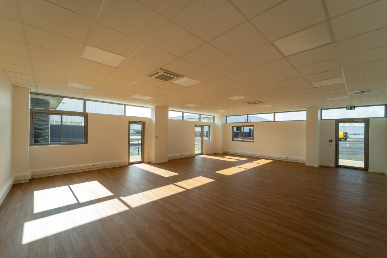 Une pièce spacieuse et vide avec de grandes fenêtres, des murs blancs, un plafond suspendu et un plancher en bois, éclairée par la lumière du soleil qui passe à travers les fenêtres.