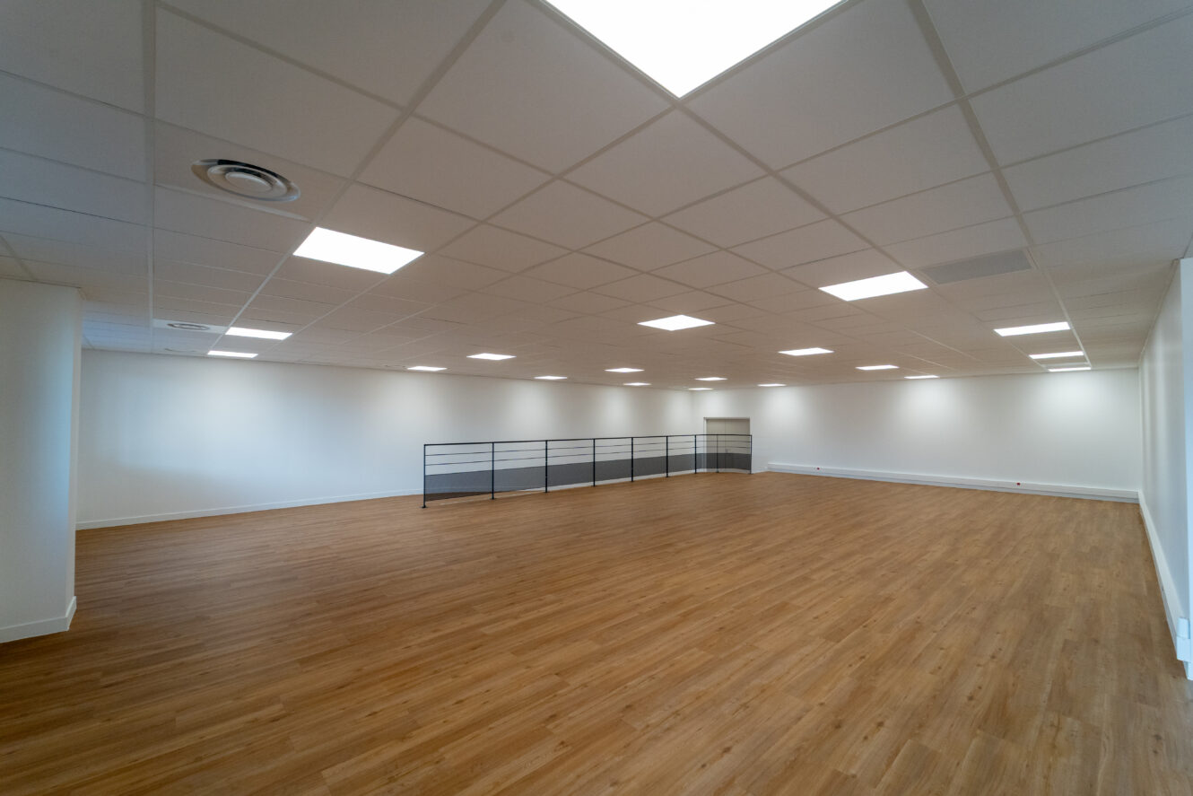 Grande pièce vide avec un sol en bois, des murs blancs, un plafond grillagé avec un éclairage fluorescent et une rampe métallique sur un côté.