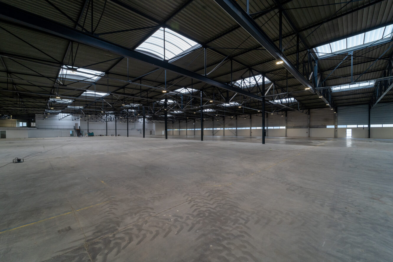 Vue large d'un entrepôt industriel vide avec de hauts plafonds, des poutres apparentes, un sol en béton, des puits de lumière et un éclairage minimal.