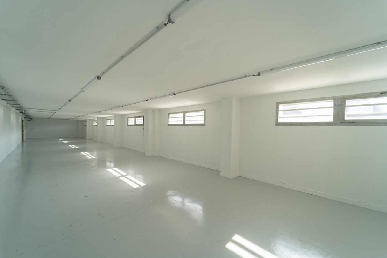 Une grande pièce vide, peinte en blanc, avec plusieurs fenêtres rectangulaires et des lampes fluorescentes au plafond. La lumière du soleil pénètre à l'intérieur, créant des reflets sur le sol.