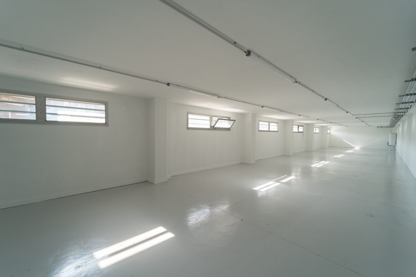 Pièce industrielle vide, peinte en blanc, avec de multiples petites fenêtres, un éclairage zénithal et un sol lisse et brillant reflétant la lumière naturelle.