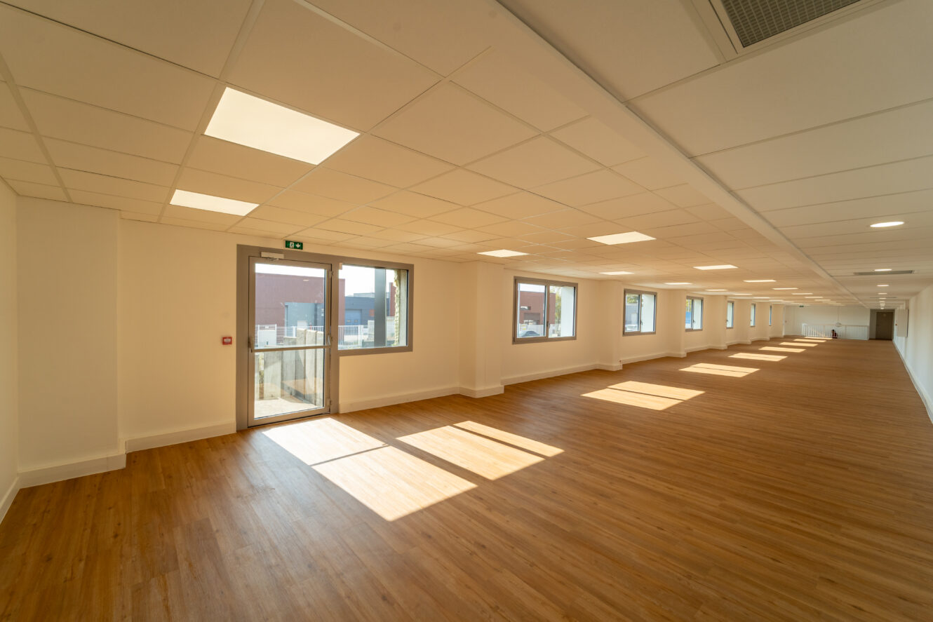 Pièce spacieuse et vide avec un sol en bois, des murs blancs, de grandes fenêtres sur un côté et des plafonniers, avec une seule porte et une fenêtre donnant sur l'extérieur.