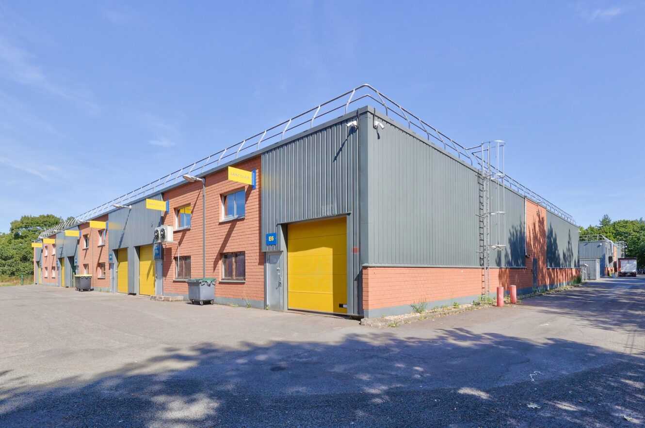 Une rangée d'entrepôts industriels avec des extérieurs en brique et en métal, chacun doté d'une porte de garage jaune vif et d'une enseigne, sous un ciel bleu clair.