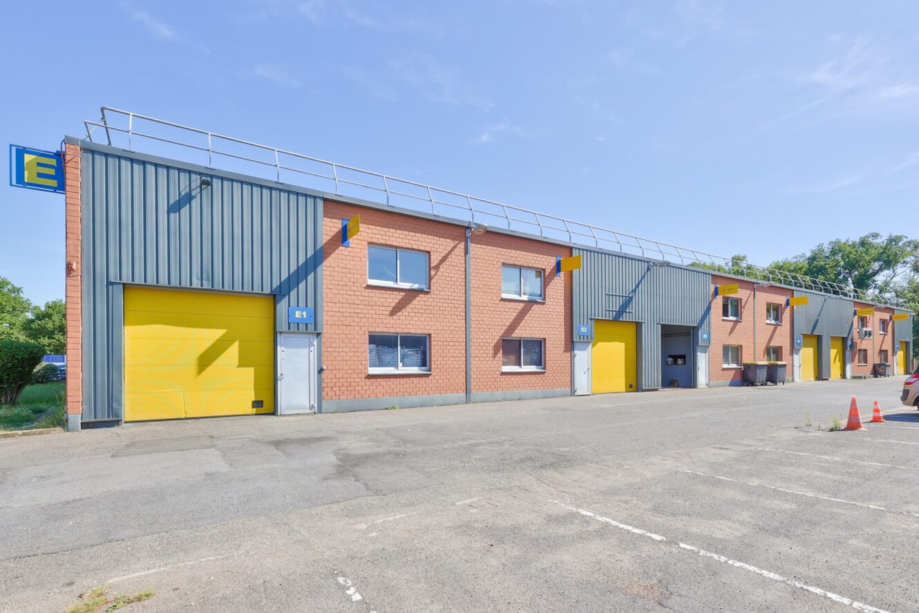 Une rangée d'entrepôts industriels avec des portes roulantes jaunes et des façades en briques, sous un ciel clair, à côté d'un parking vide.