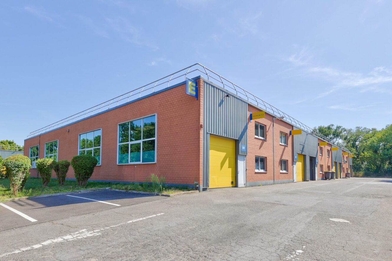 Un entrepôt industriel moderne avec des murs en briques rouges, de grandes fenêtres et des portes jaunes, situé à côté d'un parking vide sous un ciel bleu clair.