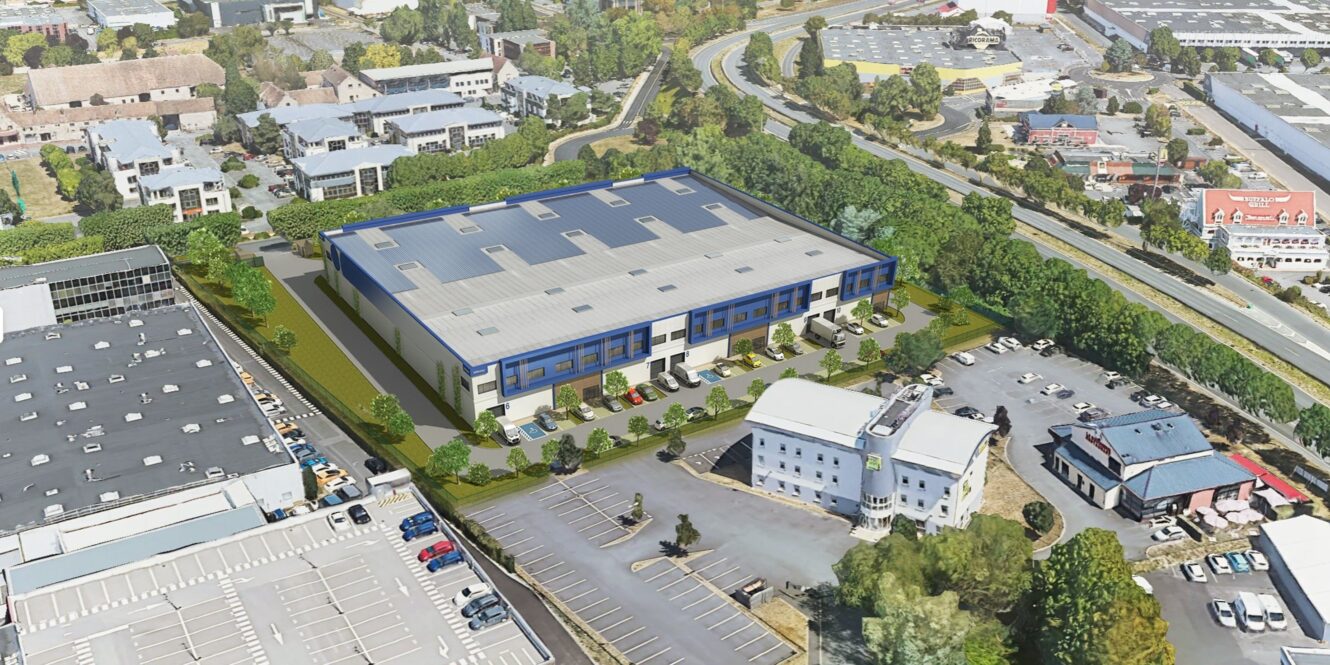 Vue aérienne d'un grand entrepôt au toit bleu entouré de parkings, d'arbres et d'immeubles de bureaux voisins dans une zone commerciale.