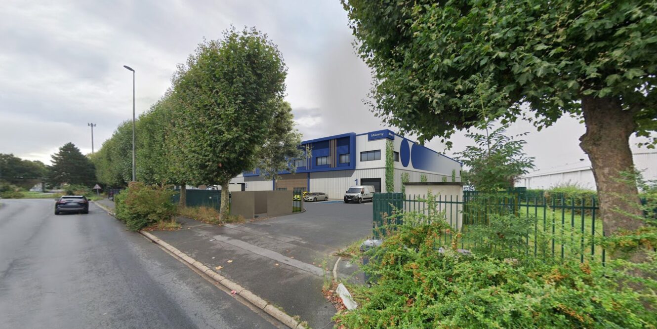 Un bâtiment industriel moderne aux accents bleus avec un quai de chargement se trouve derrière une zone clôturée le long d'une rue bordée d'arbres ; des voitures sont garées à proximité sous un ciel nuageux.