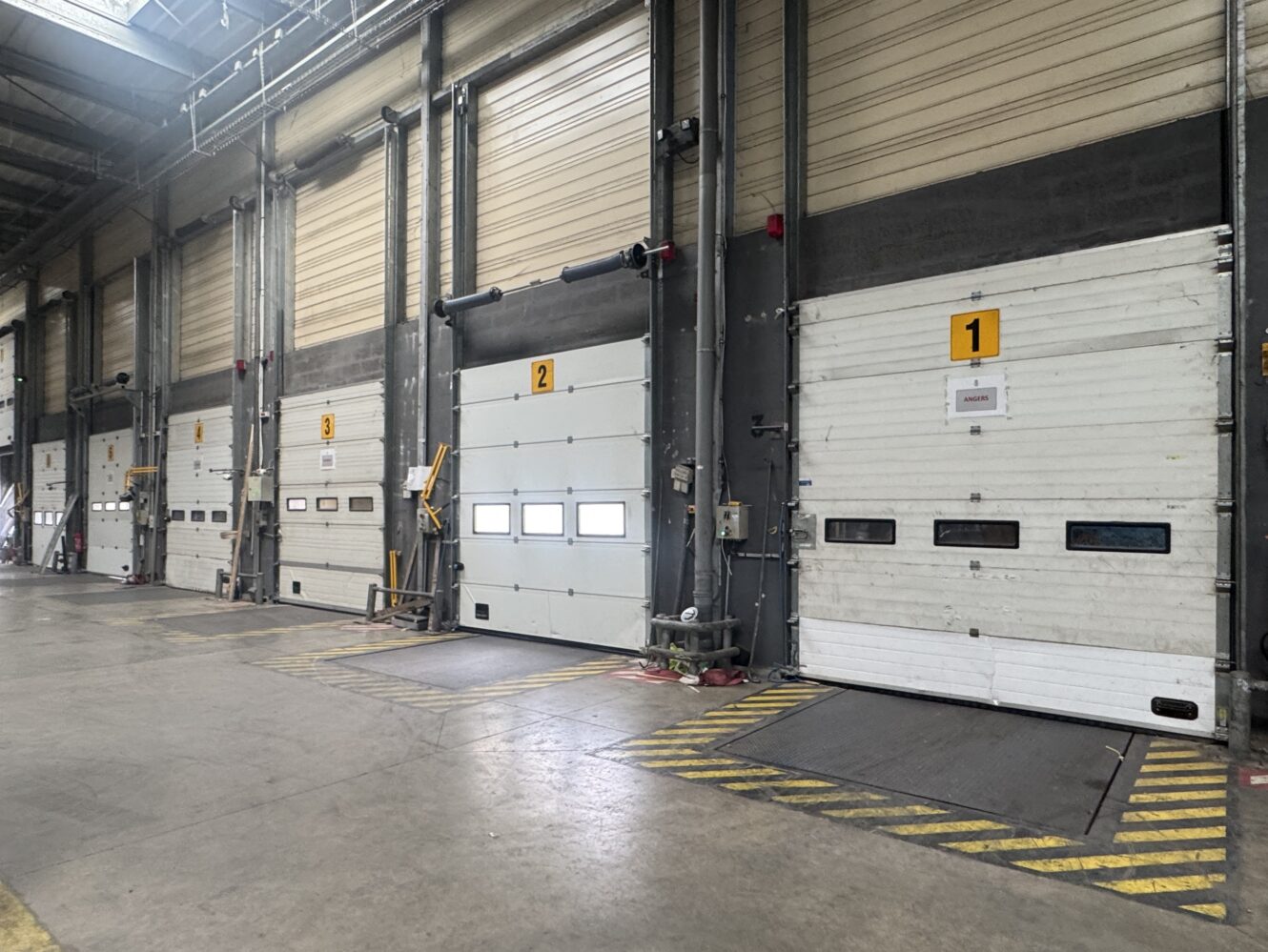Quatre portes de quai de chargement industriel numérotées 1 et 2 à l'intérieur d'un entrepôt ; la zone est vide et délimitée par des lignes de sécurité jaunes.