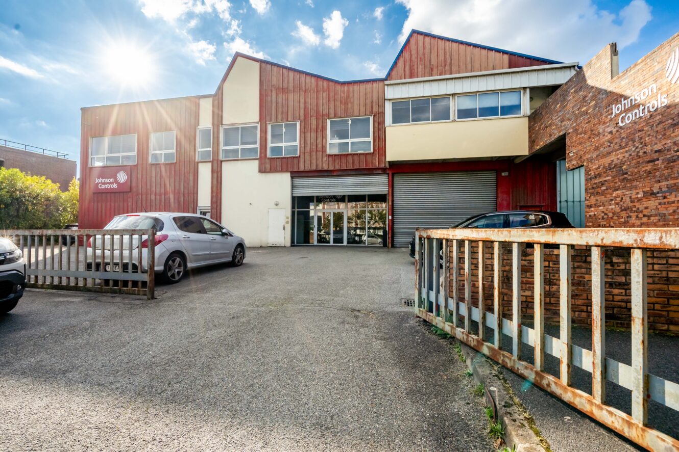 Un bâtiment industriel de deux étages avec un bardage rouge et blanc, étiqueté Johnson Controls, avec des voitures garées devant et un portail métallique partiellement ouvert.