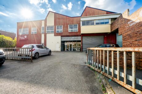 Un bâtiment industriel de deux étages avec un bardage rouge et blanc, étiqueté Johnson Controls, avec des voitures garées devant et un portail métallique partiellement ouvert.