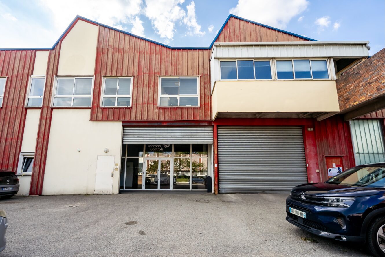 Un bâtiment de type entrepôt avec un bardage en bois rouge, de grandes fenêtres, une porte métallique à enroulement et des voitures garées devant.