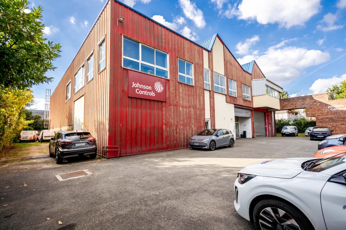 Un bâtiment industriel rouge avec une enseigne Johnson Controls, des voitures garées devant, et des arbres sur la gauche sous un ciel partiellement nuageux.