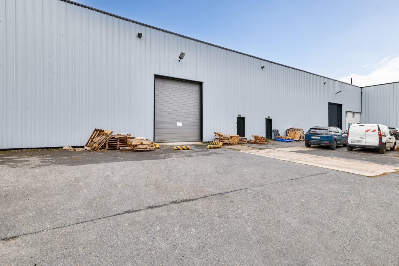 L'extérieur d'un entrepôt industriel gris avec une porte roulante fermée, des palettes en bois empilées, deux voitures garées et une camionnette blanche sur un terrain pavé sous un ciel dégagé.