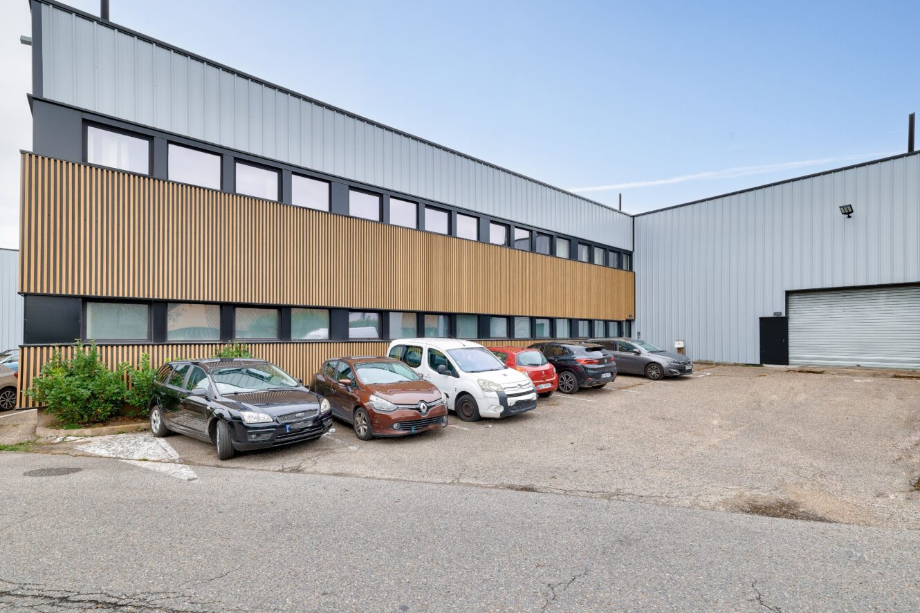 Plusieurs voitures sont garées devant un bâtiment industriel moderne à la façade grise et aux panneaux de bois.