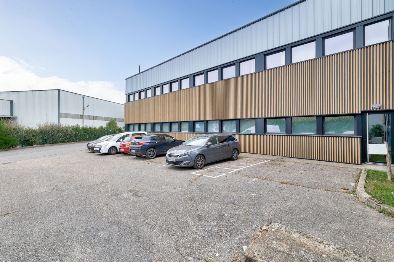 Par temps clair, quelques voitures sont garées devant un bâtiment industriel ou de bureaux moderne avec un bardage en bois et en métal.