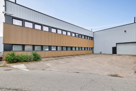 Bâtiment industriel moderne doté d'un revêtement métallique, de fenêtres horizontales, de lambris décoratifs en bois et d'un parking pavé vide à l'avant, photographié de jour sous un ciel dégagé.