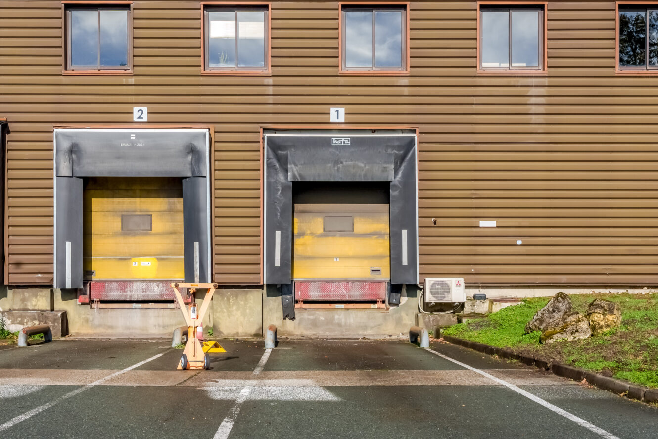 Deux quais de chargement avec des portes jaunes sont fermés ; l'un est étiqueté "1" et l'autre "2". Une barrière en bois bloque l'accès au quai 2. Le bâtiment a un extérieur brun et plusieurs fenêtres au-dessus.