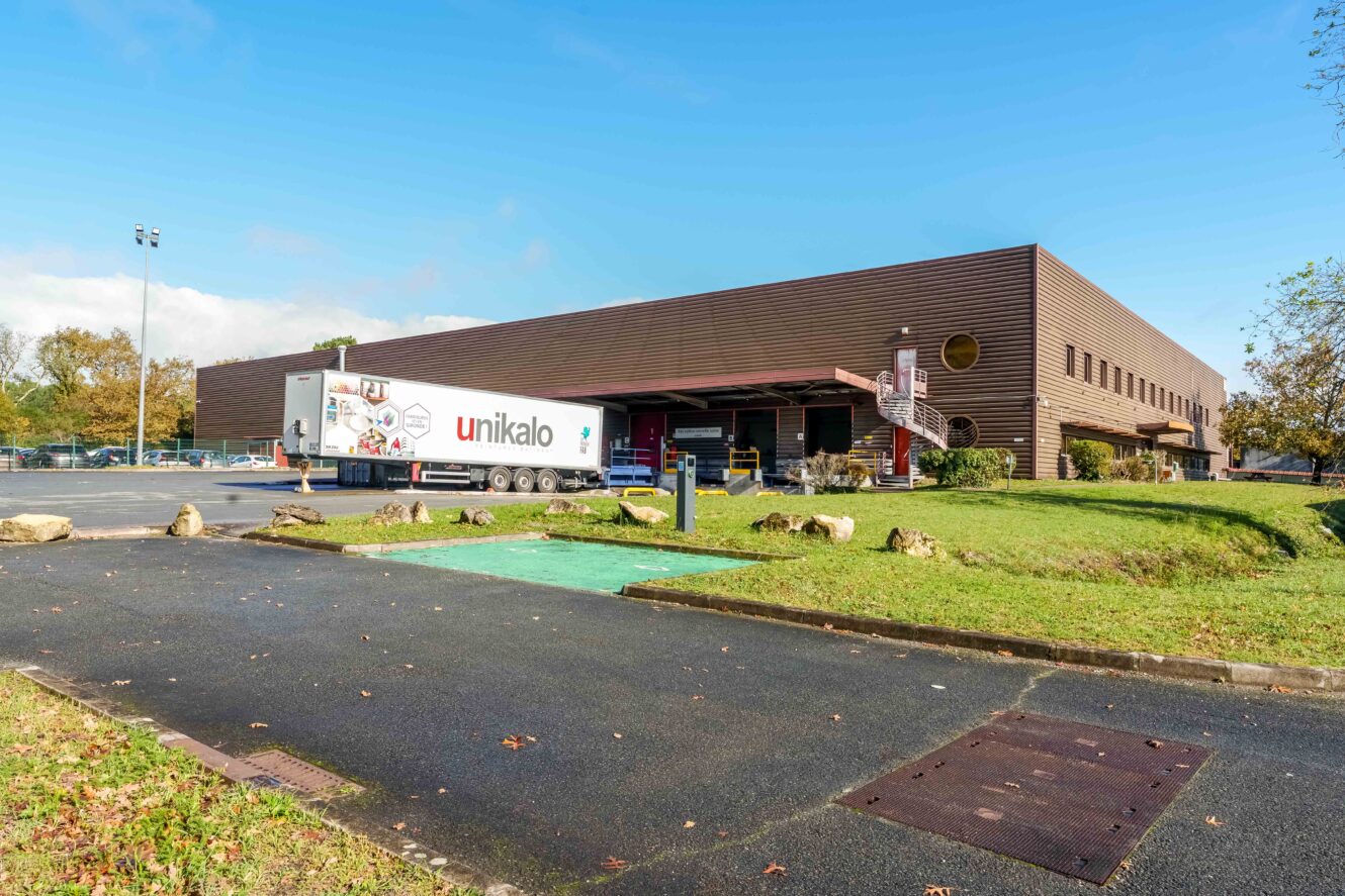 Un grand entrepôt industriel brun avec un quai de chargement, un camion remorque garé à l'extérieur et des zones herbeuses au premier plan.