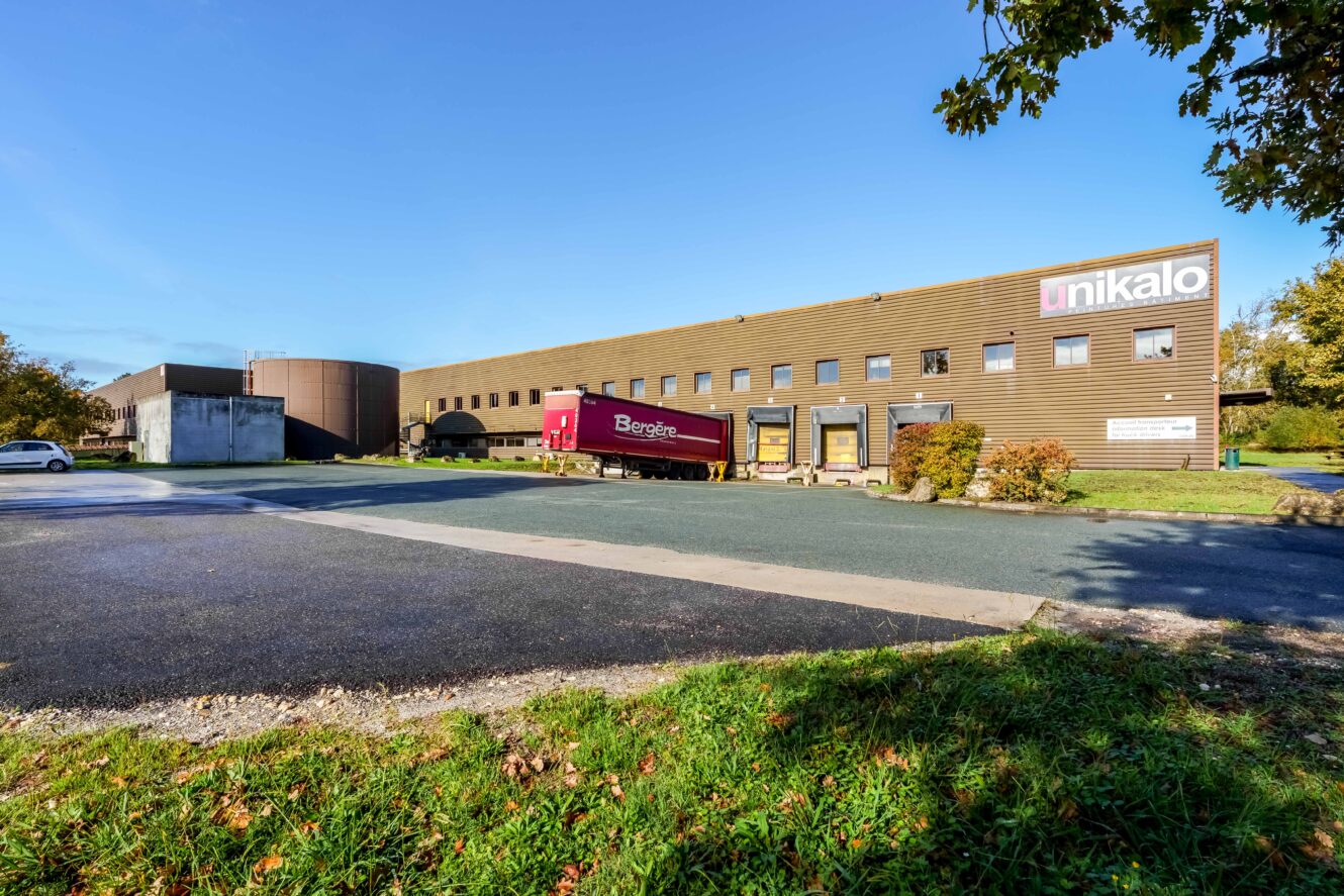 Un grand bâtiment industriel avec un panneau "unikalo", un camion de livraison rouge garé à l'extérieur et un ciel bleu clair au-dessus de la tête.