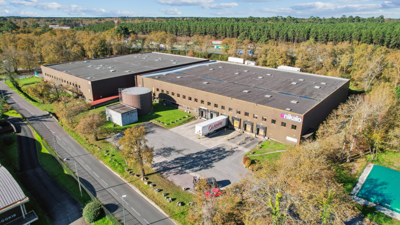 Vue aérienne d'un grand entrepôt industriel brun avec des quais de chargement, un camion garé à l'extérieur et entouré d'arbres et de routes.