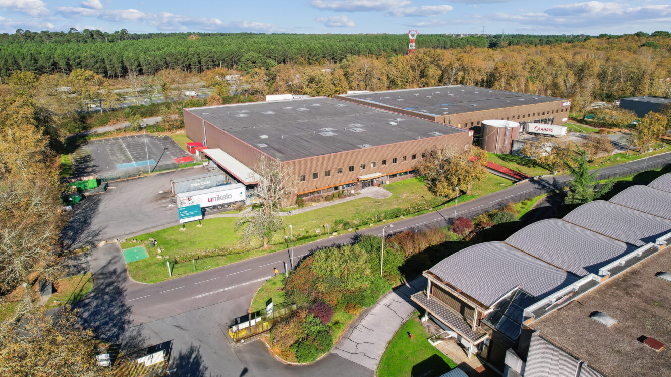 Vue aérienne d'un grand complexe industriel avec parking adjacent, entouré d'arbres et de verdure, sous un ciel partiellement nuageux.