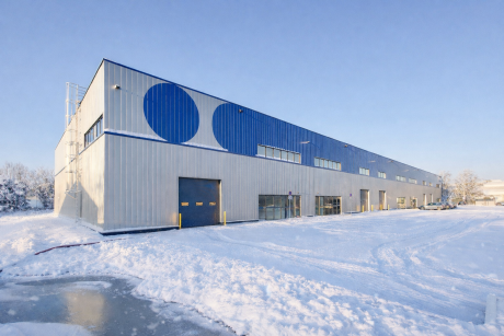 Un grand entrepôt industriel avec des accents bleus et des motifs circulaires sur le mur supérieur, entouré de neige et d'un ciel d'hiver dégagé.