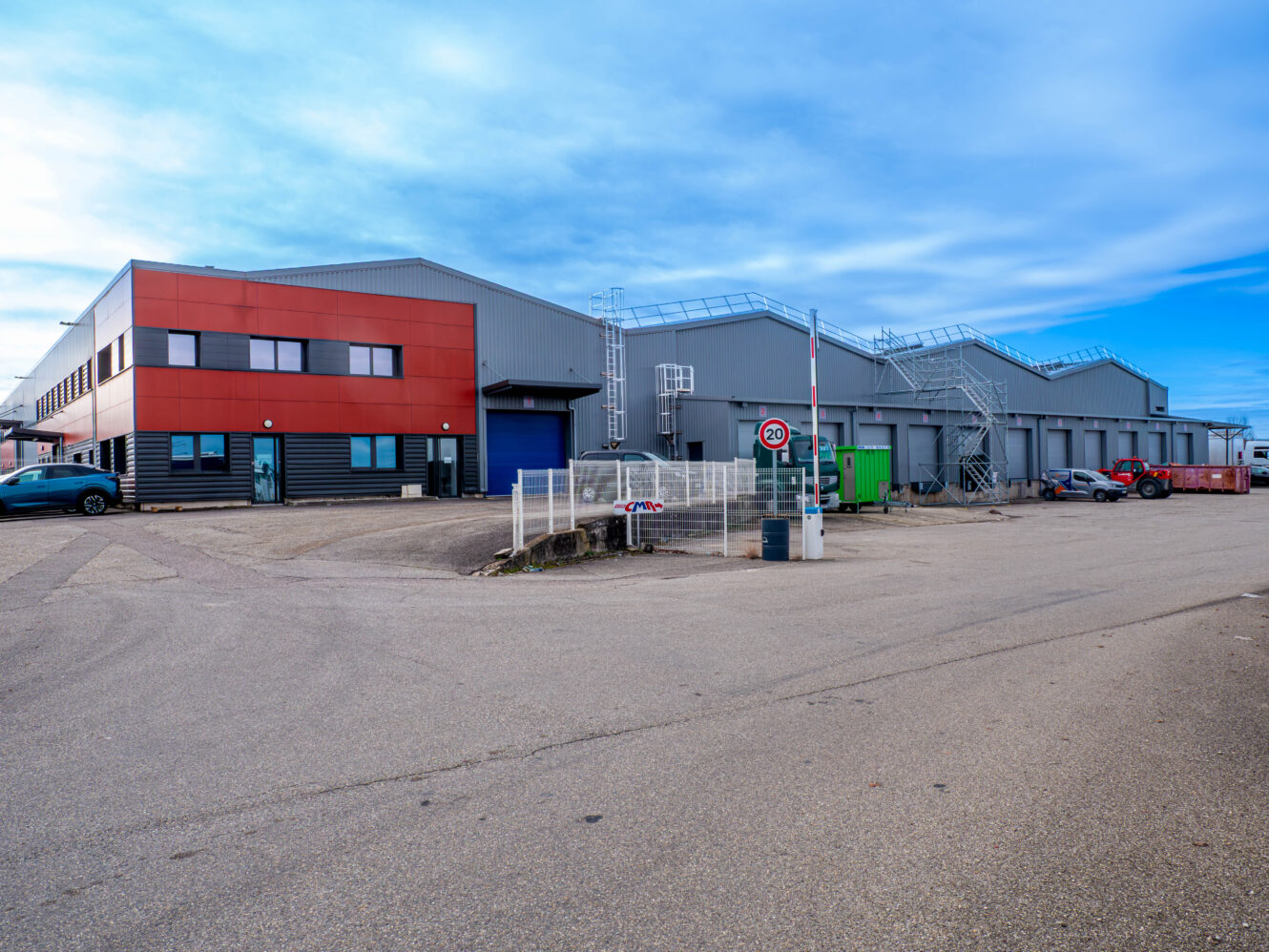 Un grand entrepôt industriel avec un extérieur rouge et gris, plusieurs quais de chargement, des escaliers métalliques et une porte d'entrée, sous un ciel partiellement nuageux.