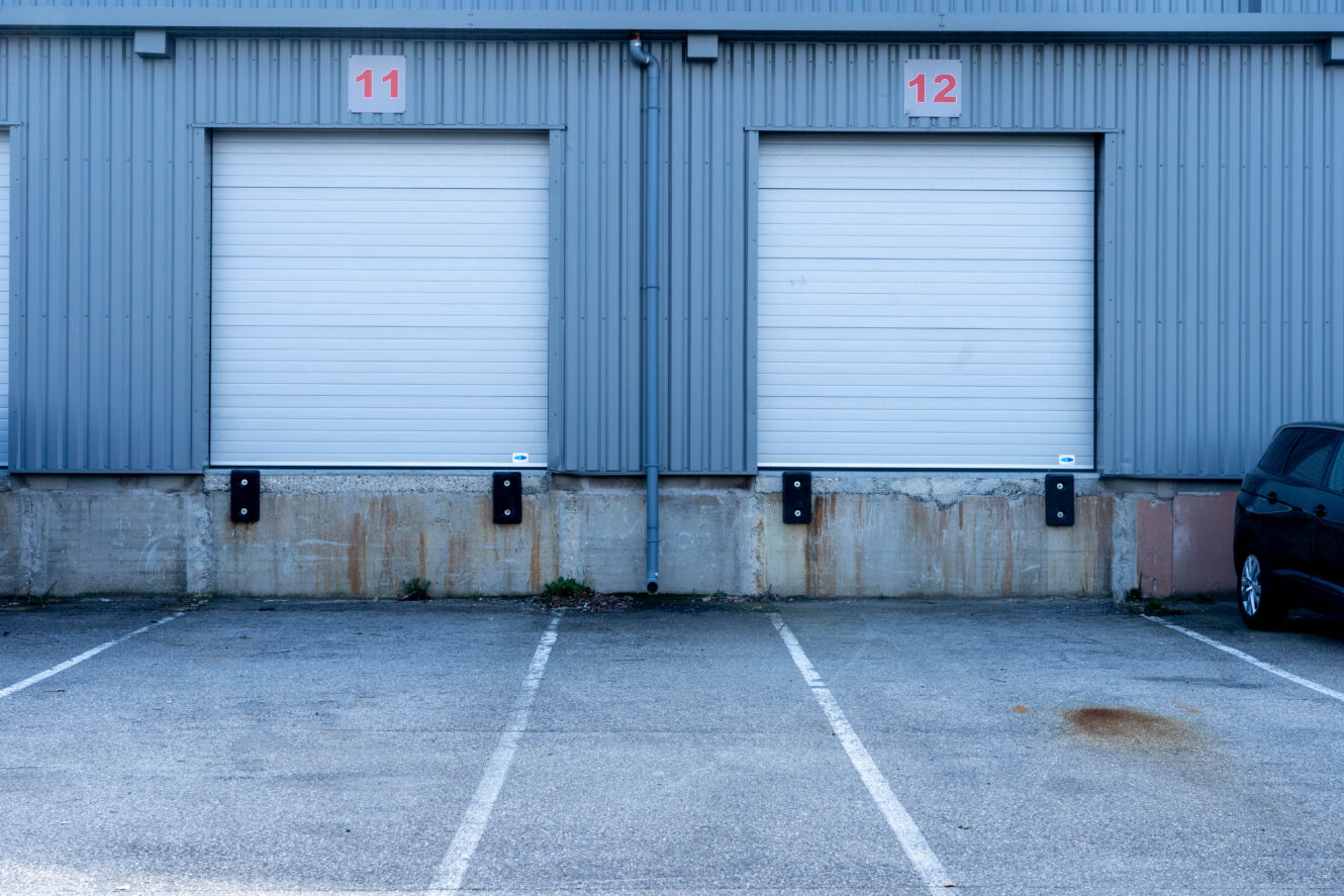 Deux portes de quai de chargement fermées, numérotées 11 et 12, sur un bâtiment industriel gris, avec des places de parking vides devant et une partie d'une voiture noire visible sur la droite.