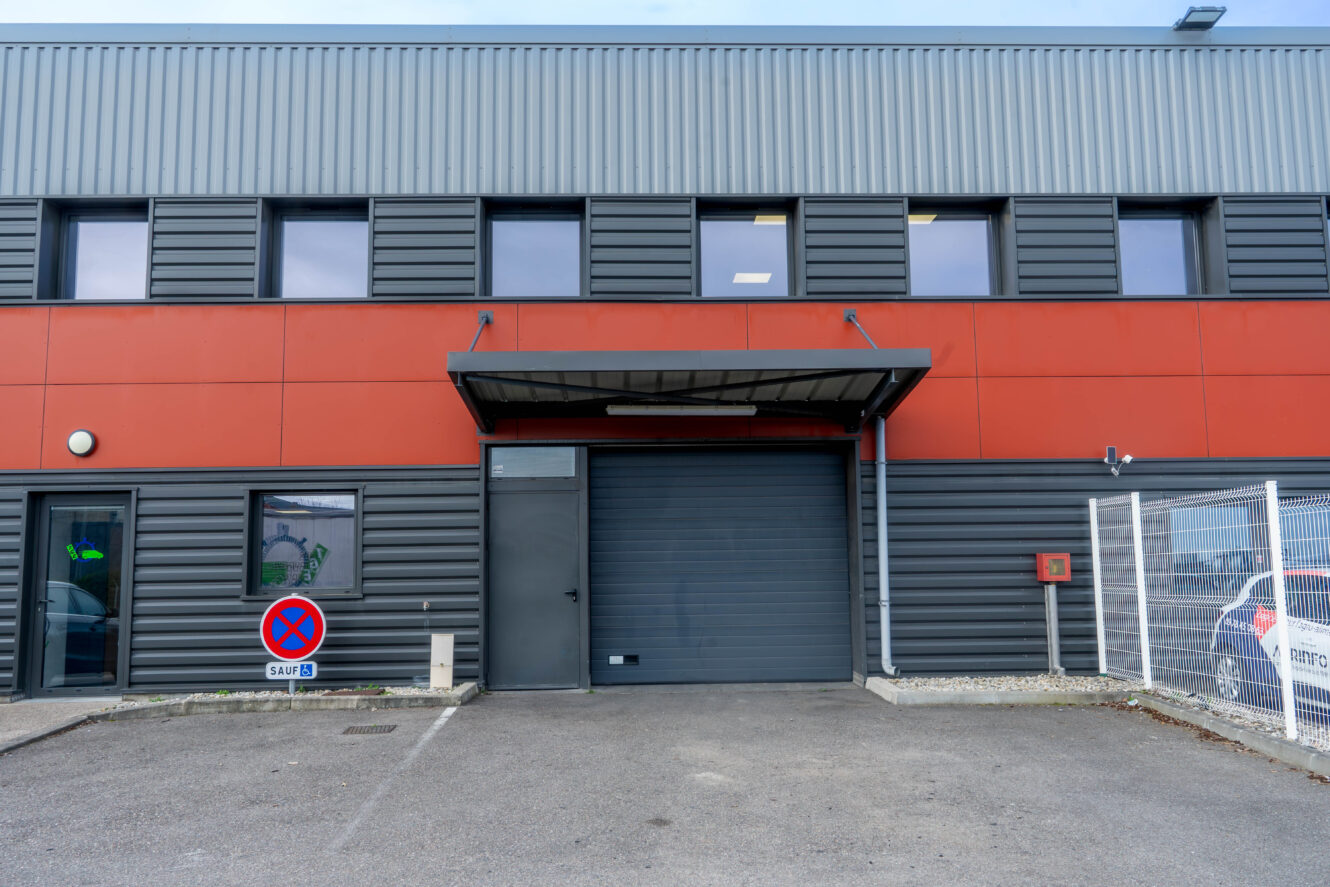 Un bâtiment industriel moderne avec une porte de garage noire fermée, une porte grise, un panneau d'interdiction de stationner et une zone clôturée sur la droite.