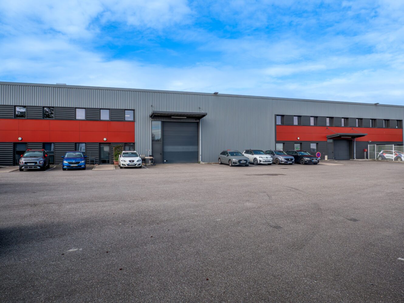 Grand bâtiment industriel à la façade grise et rouge, plusieurs voitures garées devant, et un large terrain asphalté sous un ciel partiellement nuageux.