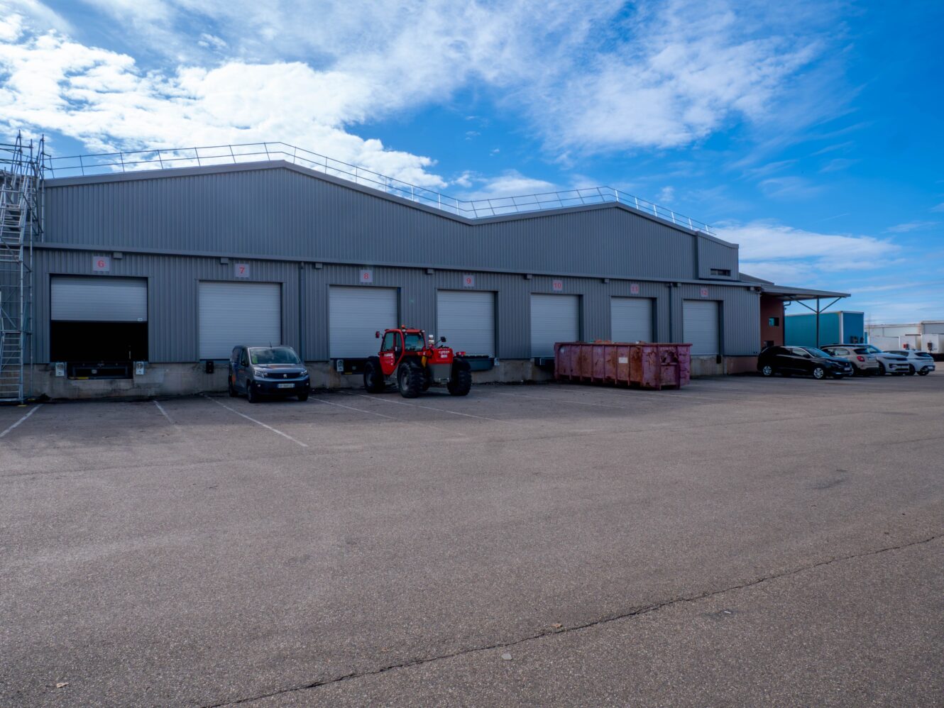 Un grand entrepôt industriel avec plusieurs quais de chargement, des voitures garées, un tracteur rouge et une benne à ordures rose dans le parking sous un ciel bleu.