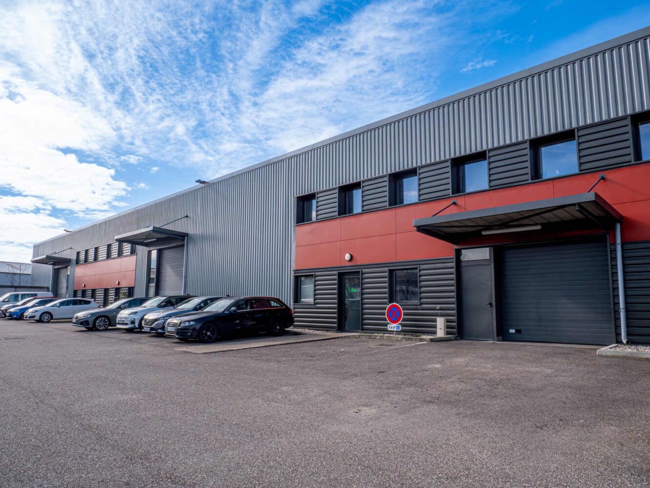 Une rangée de voitures garées devant un entrepôt industriel moderne à l'extérieur gris et rouge, sous un ciel partiellement nuageux.