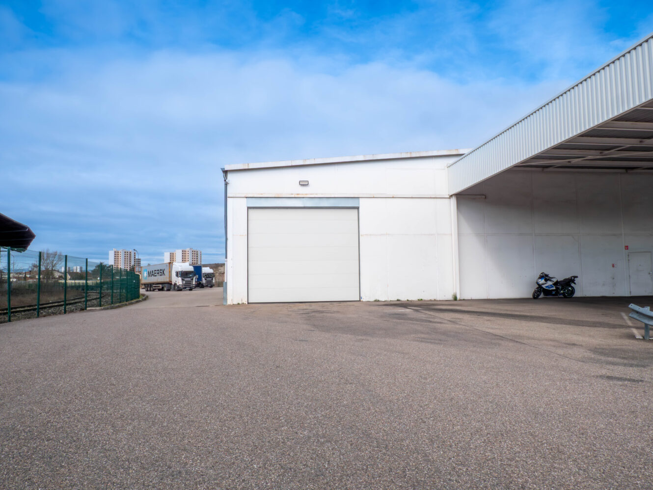 Grand entrepôt blanc avec une porte de garage fermée, une moto garée près du mur et un camion de livraison en arrière-plan sous un ciel partiellement nuageux.