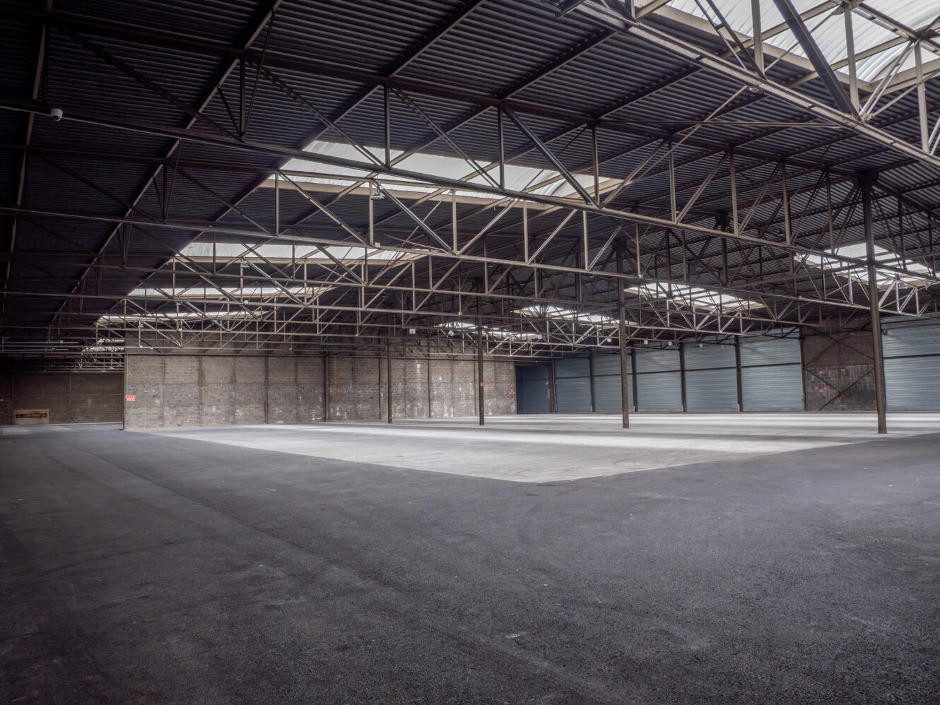 Grand entrepôt vide avec un sol en béton, un haut plafond métallique, des poutres apparentes et des murs en briques. La lumière naturelle pénètre par les fenêtres du toit.