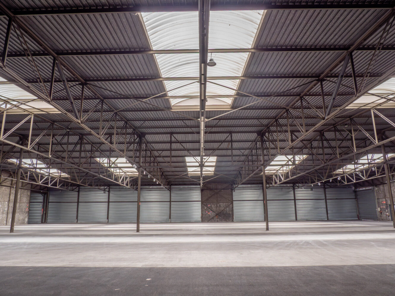 Intérieur d'un entrepôt industriel vide avec des poutres métalliques apparentes, un sol en béton, un haut plafond et de grandes portes métalliques fermées bordant le mur du fond. La lumière naturelle pénètre par des puits de lumière.
