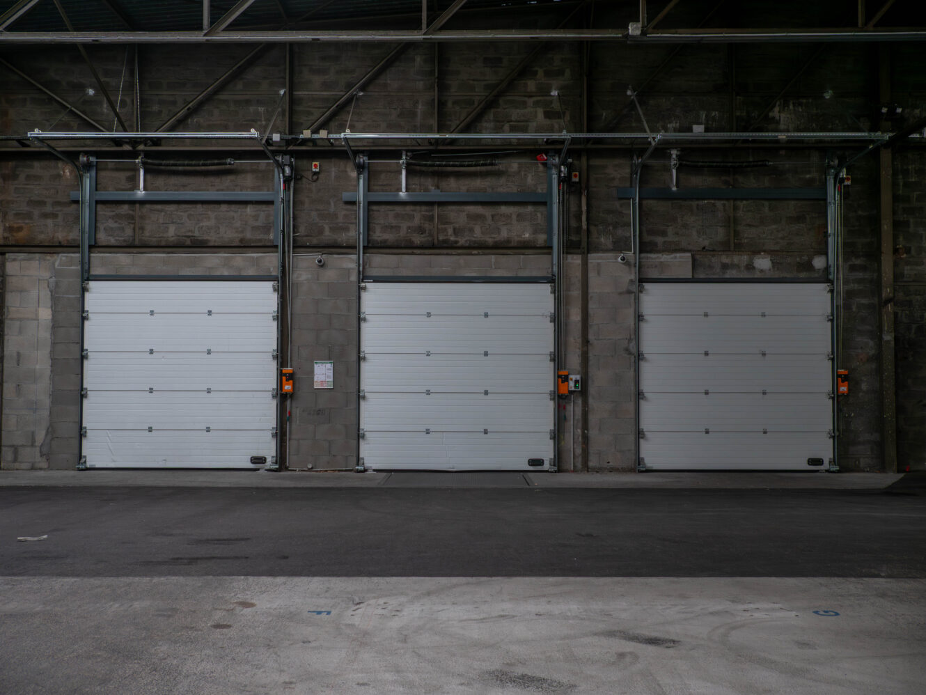 Trois portes de garage industrielles fermées, encastrées dans un mur de béton gris, chacune avec un moteur d'ouverture et un panneau de commande orange monté à côté.