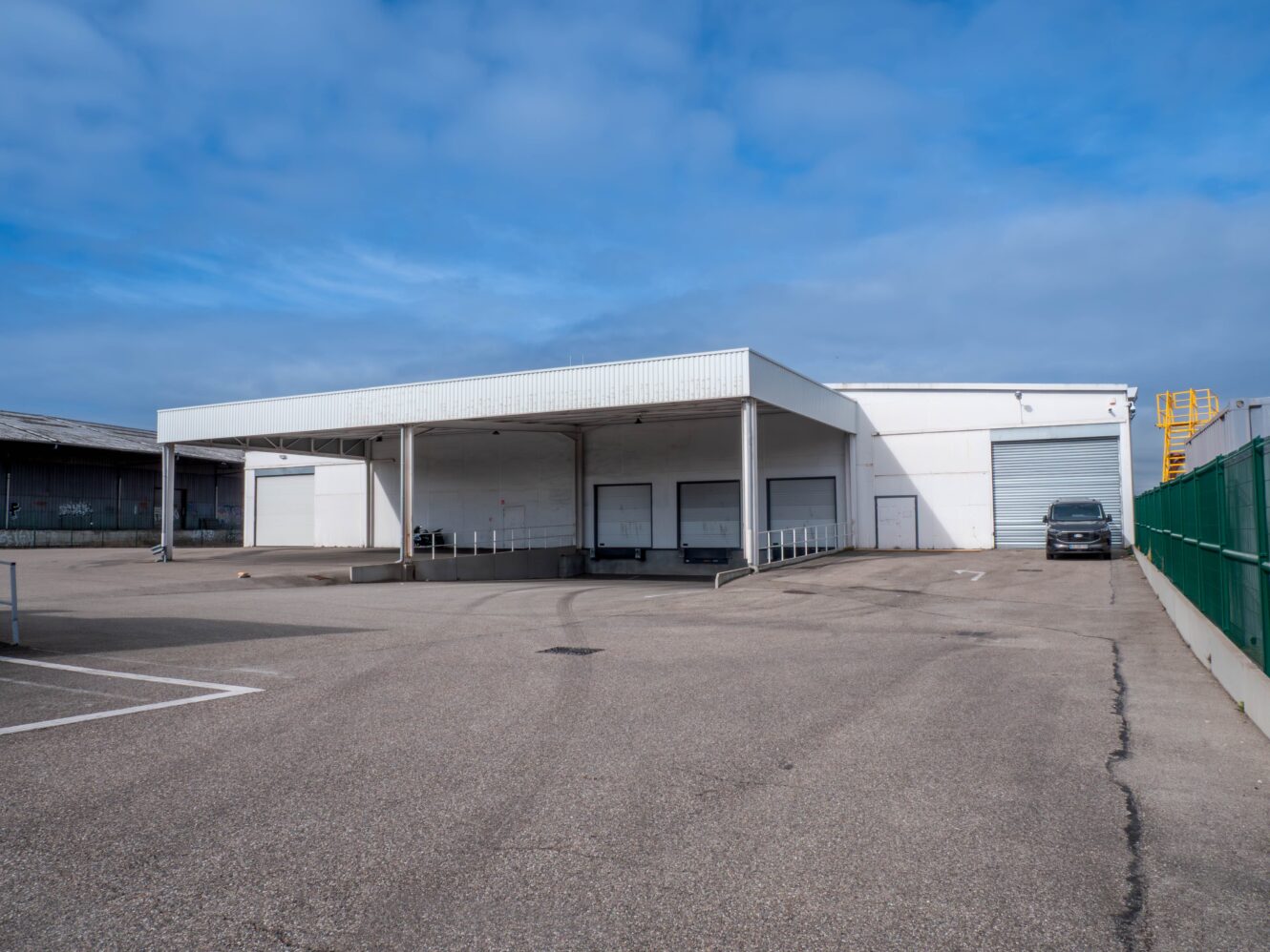 Un grand entrepôt industriel blanc avec plusieurs quais de chargement, une zone couverte et un parking vide sous un ciel partiellement nuageux.