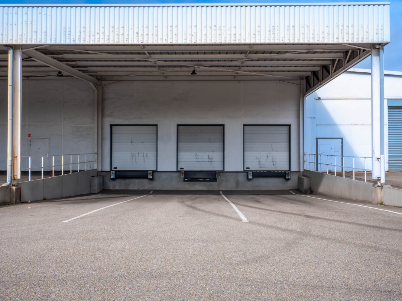 Quai de chargement vide avec trois portes de quai fermées et des places de stationnement marquées devant un entrepôt.