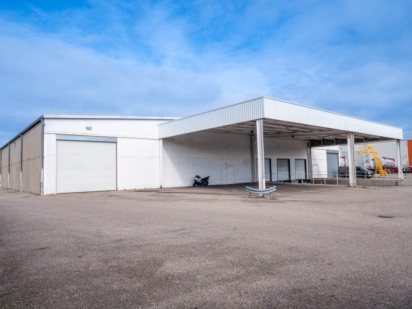 Un grand entrepôt industriel blanc avec une zone de chargement couverte, des portes de garage fermées et une moto garée près du mur sous un ciel nuageux.