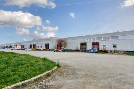 Un grand entrepôt industriel avec plusieurs quais de chargement, plusieurs voitures et camions garés sous un ciel partiellement nuageux.