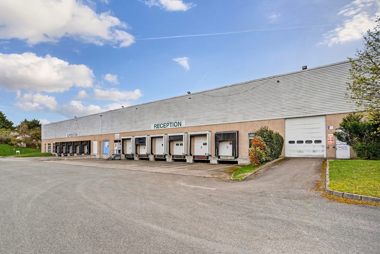 Entrepôt industriel de plain-pied doté de plusieurs quais de chargement portant l'inscription « Réception », d'une allée pavée, d'espaces verts et d'un ciel partiellement nuageux.