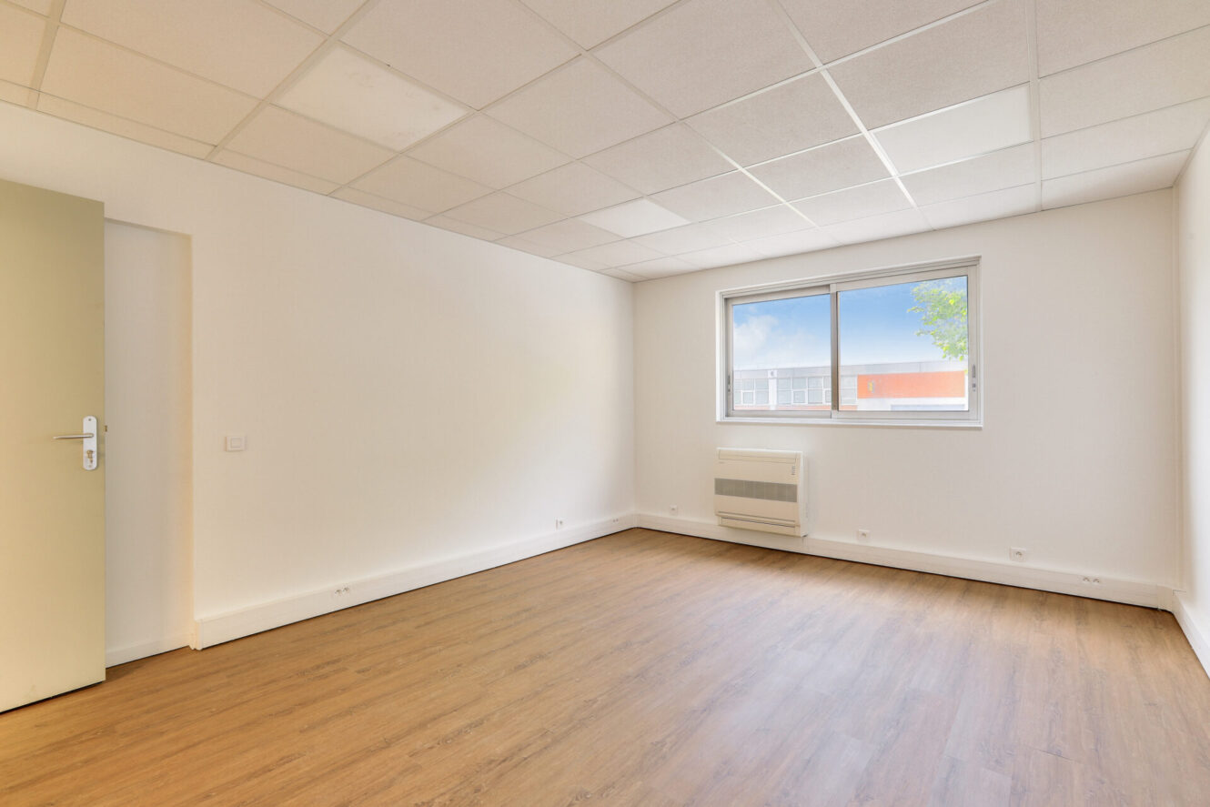 Pièce vide avec des murs blancs, un sol en bois, une fenêtre donnant sur l'extérieur, des dalles de plafond et une porte lumineuse fermée sur la gauche.
