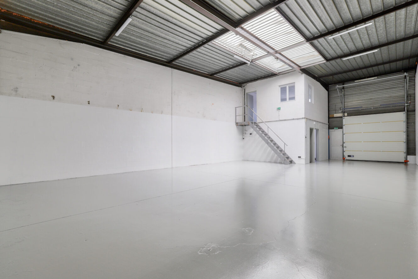 Entrepôt industriel vide avec des murs blancs, un sol en béton gris, un plafond métallique, un bureau surélevé, des escaliers et une porte roulante fermée.