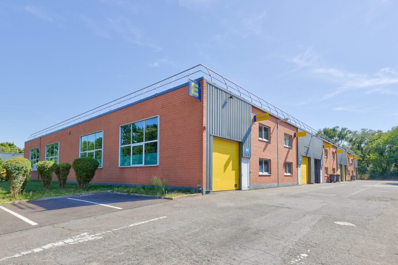 Une rangée d'entrepôts industriels avec des murs en briques rouges, de grandes fenêtres et des portes de garage jaunes près d'un parking vide.