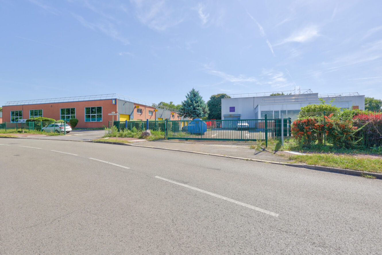 Un complexe industriel ou de bureaux clôturé avec un parking, de la verdure et une route à deux voies devant, sous un ciel bleu clair.
