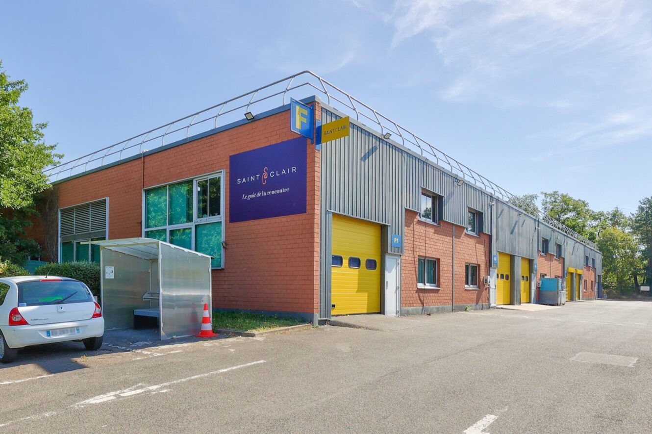 Un bâtiment industriel de deux étages avec des portes de garage jaunes, une enseigne "Saint Clair" et une voiture blanche garée devant par une journée ensoleillée.